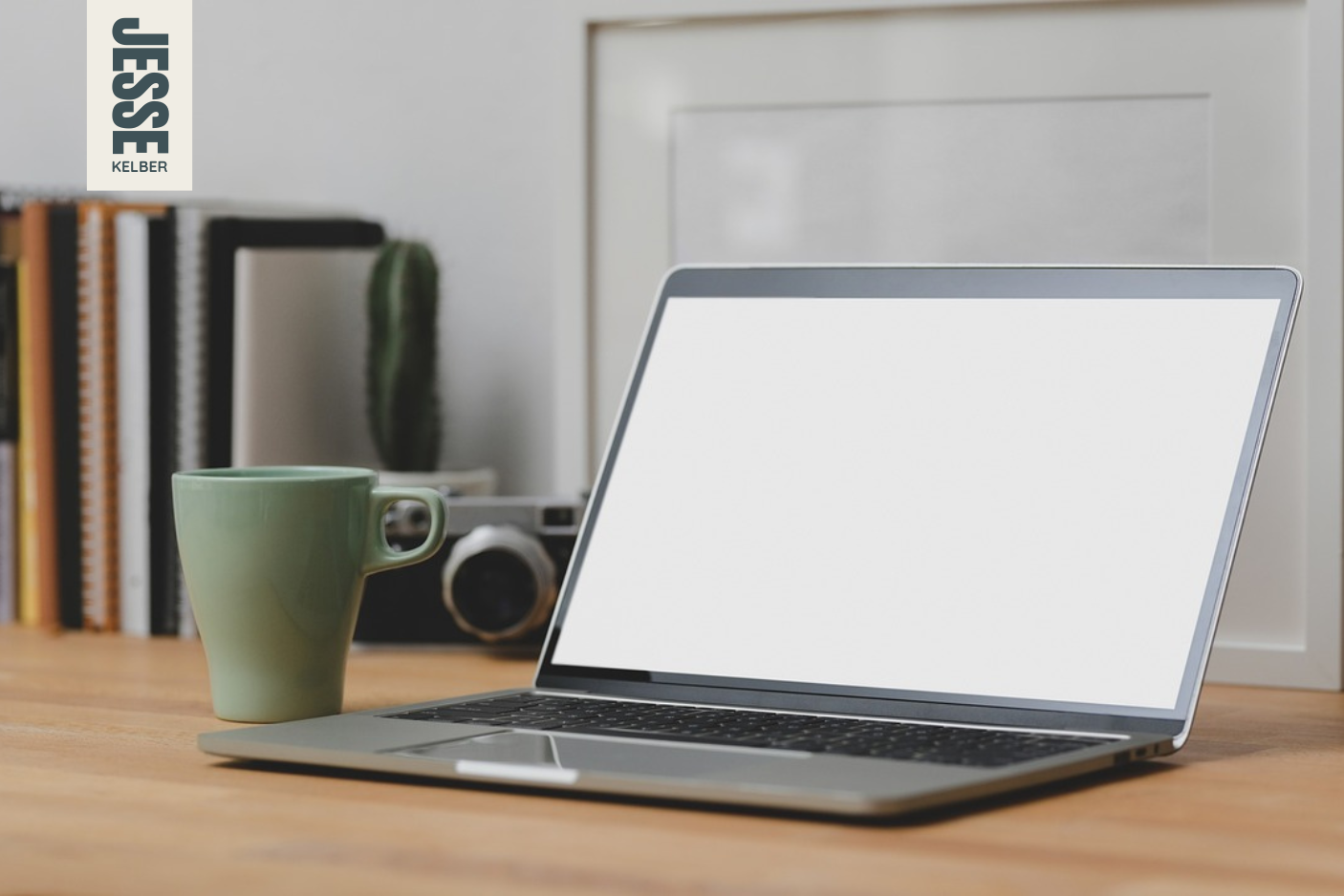 A laptop sits on a wood desk with a cup, camera, and some books in the background. The screen on the laptops is blank.